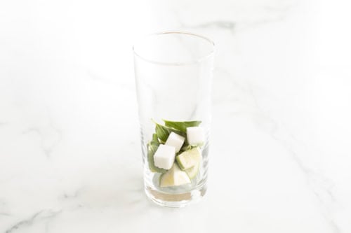 A tall glass containing lime slices, sugar cubes, mint leaves, and tequila on a marble surface.