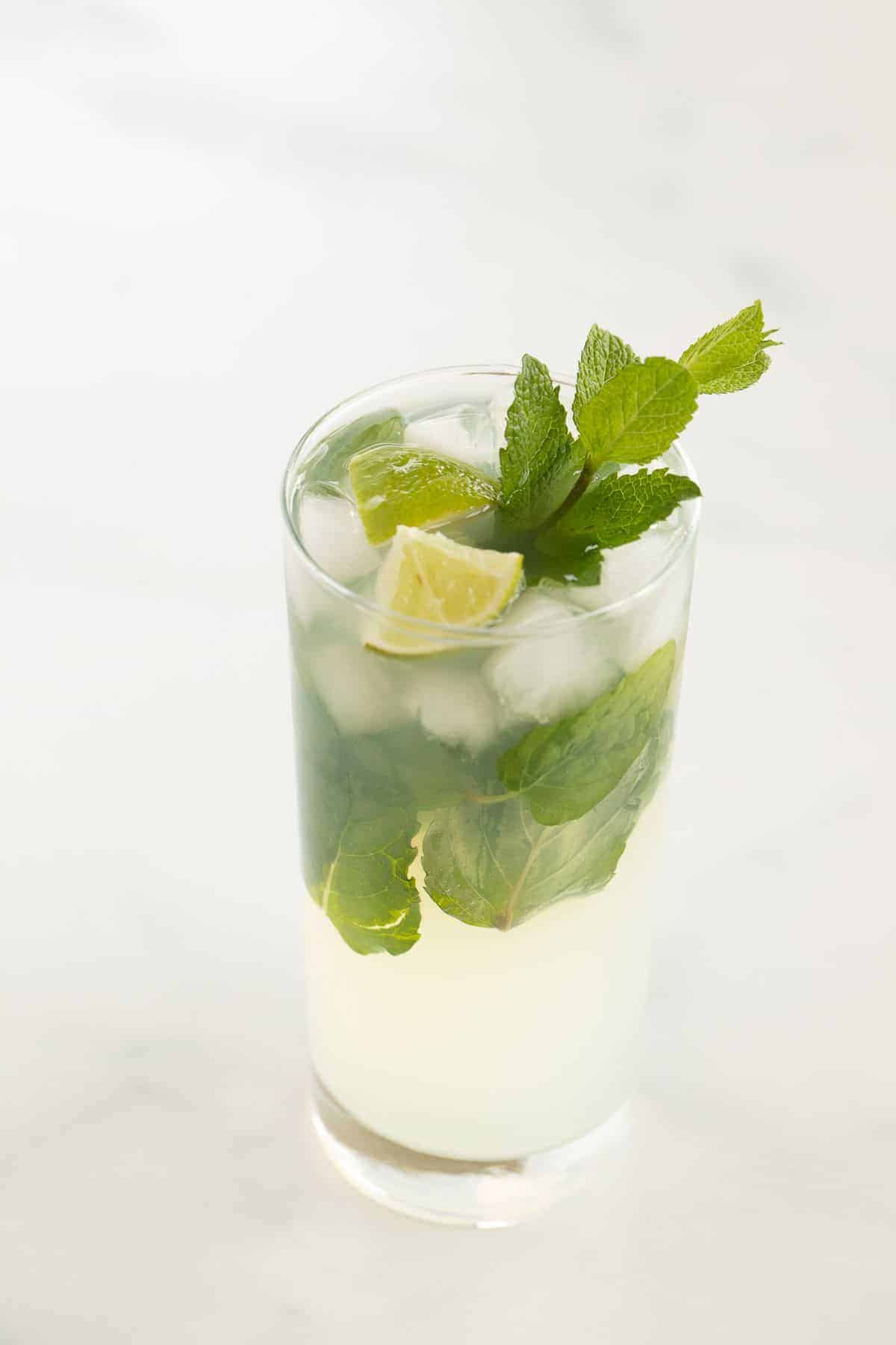 A tequila mojito in a clear glass on a white surface, garnished with lime wedges and fresh mint.