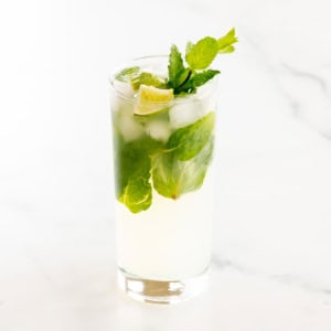 A glass of tequila mojito cocktail with lime slices and mint leaves on a marble surface.