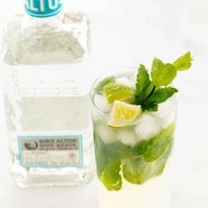 A tequila mojito in a clear glass on a white surface, garnished with lime wedges and fresh mint, bottle of tequila in the background.