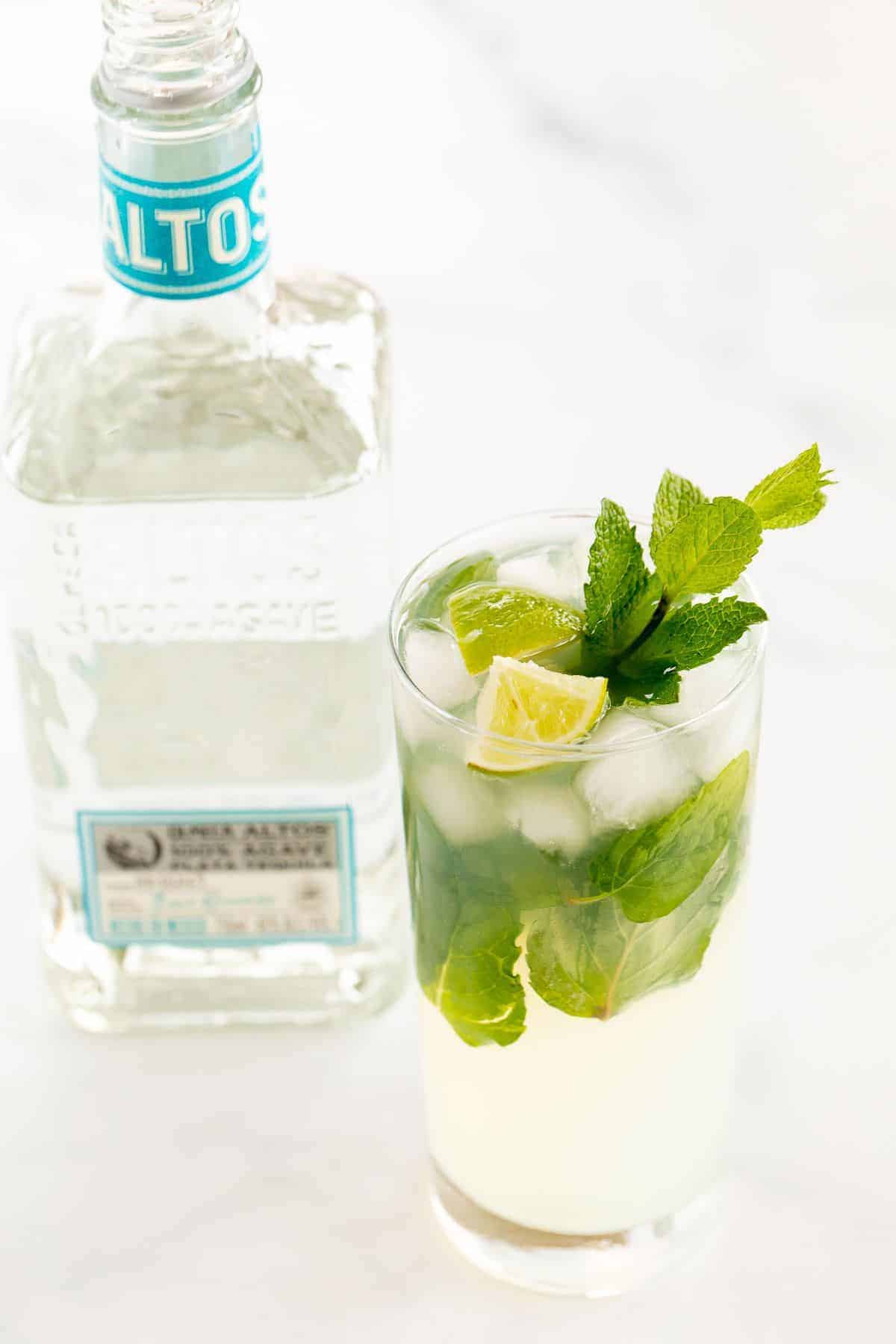 A tequila mojito in a clear glass on a white surface, garnished with lime wedges and fresh mint, bottle of tequila in the background.