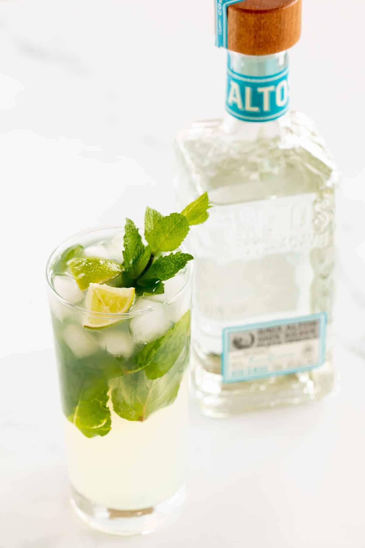 A tequila mojito in a clear glass on a white surface, garnished with lime wedges and fresh mint, bottle of tequila in the background.