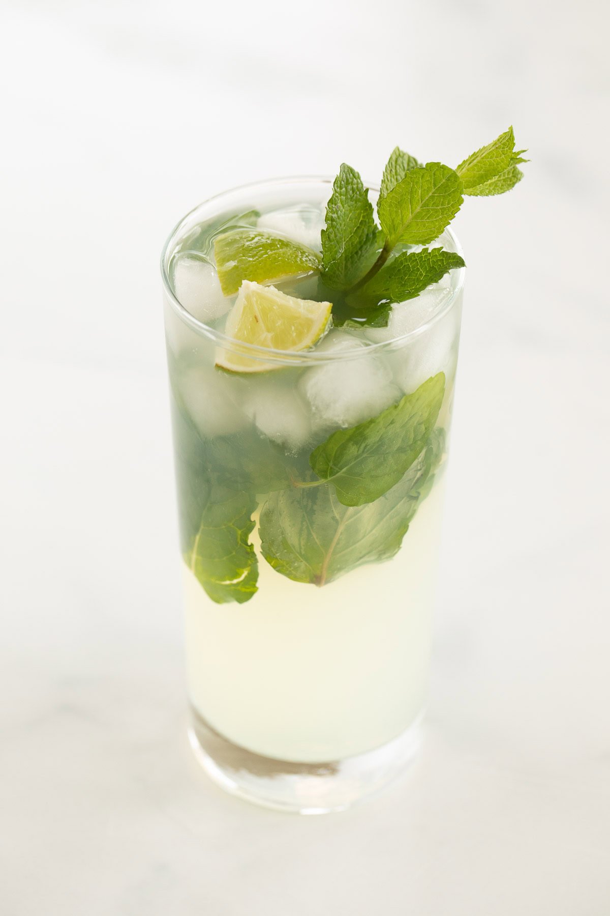 A glass of tequila mojito cocktail with lime wedges, ice cubes, and fresh mint leaves on a light background.