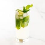 A tequila mojito in a clear glass on a white surface, garnished with lime wedges and fresh mint.
