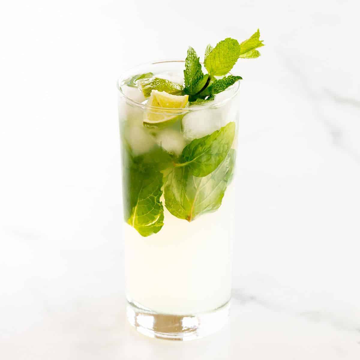 A tequila mojito in a clear glass on a white surface, garnished with lime wedges and fresh mint.