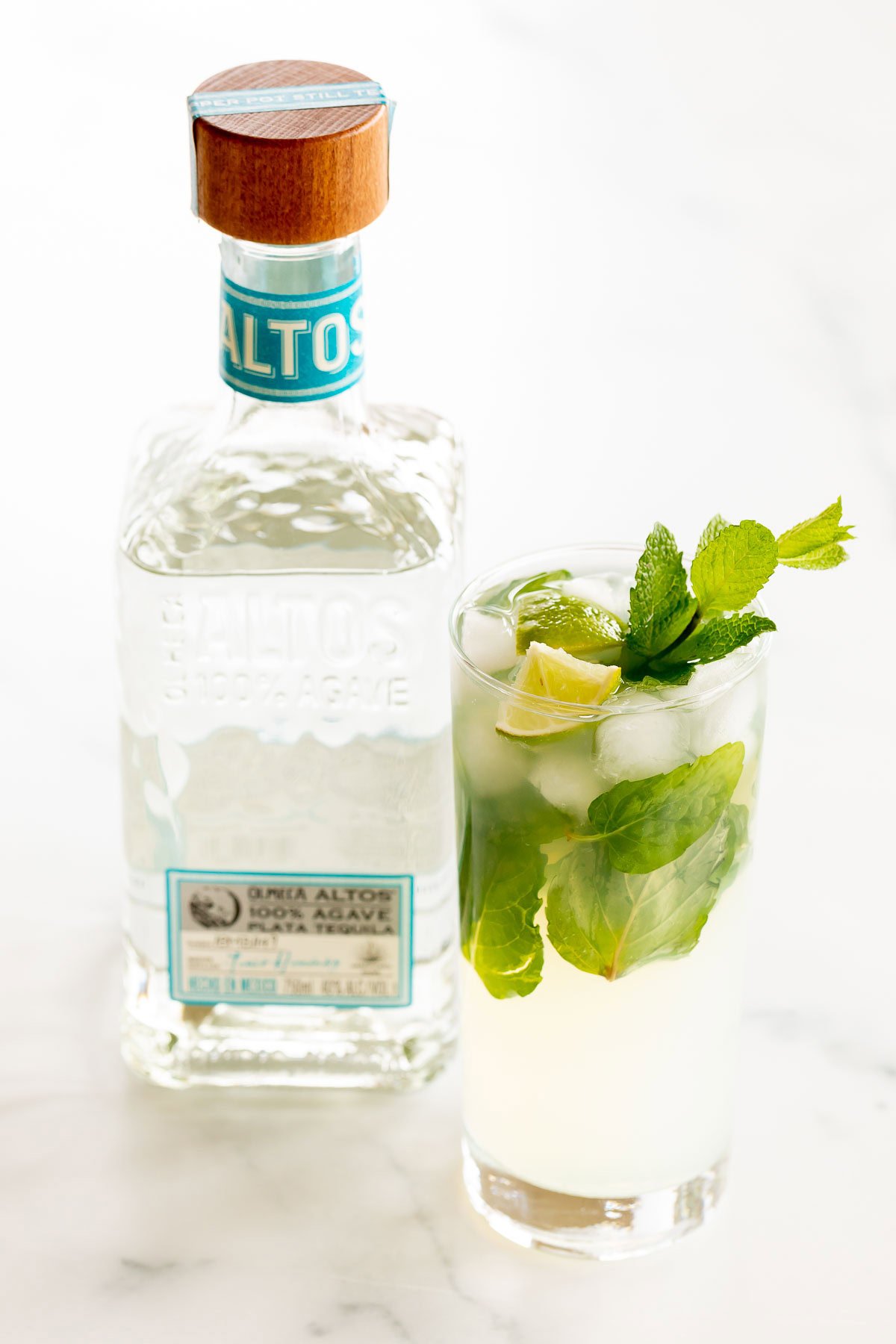 A glass of tequila mojito with lime and mint next to a bottle of Olmeca Altos tequila on a marble surface.