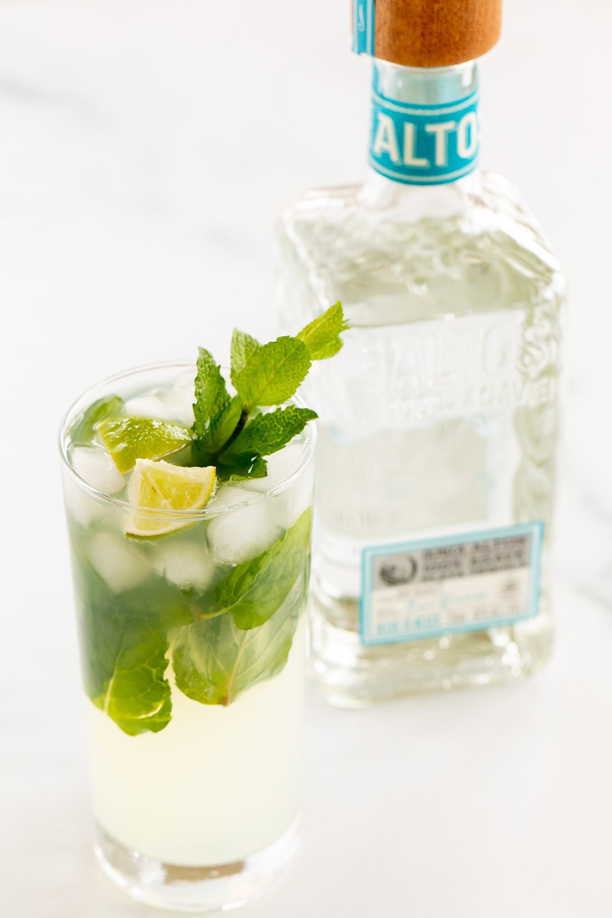 A glass of tequila mojito with lime and mint next to a bottle of altos tequila on a white surface.