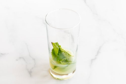 A partially filled glass with lime wedges, mint leaves, and tequila on a marble surface.