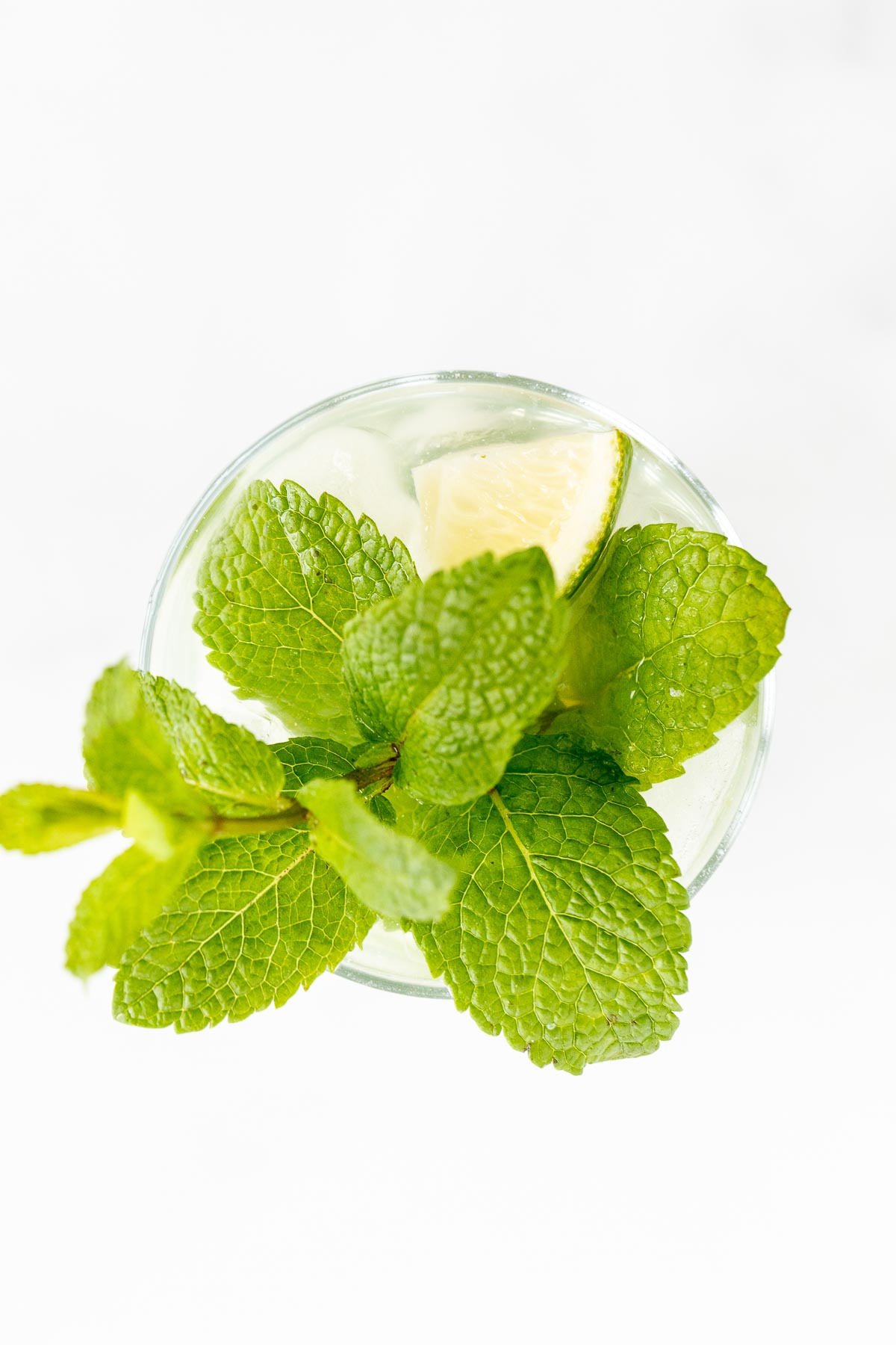 Top view of a glass of tequila mojito with ice cubes, fresh mint leaves, and a slice of lime on a white background.