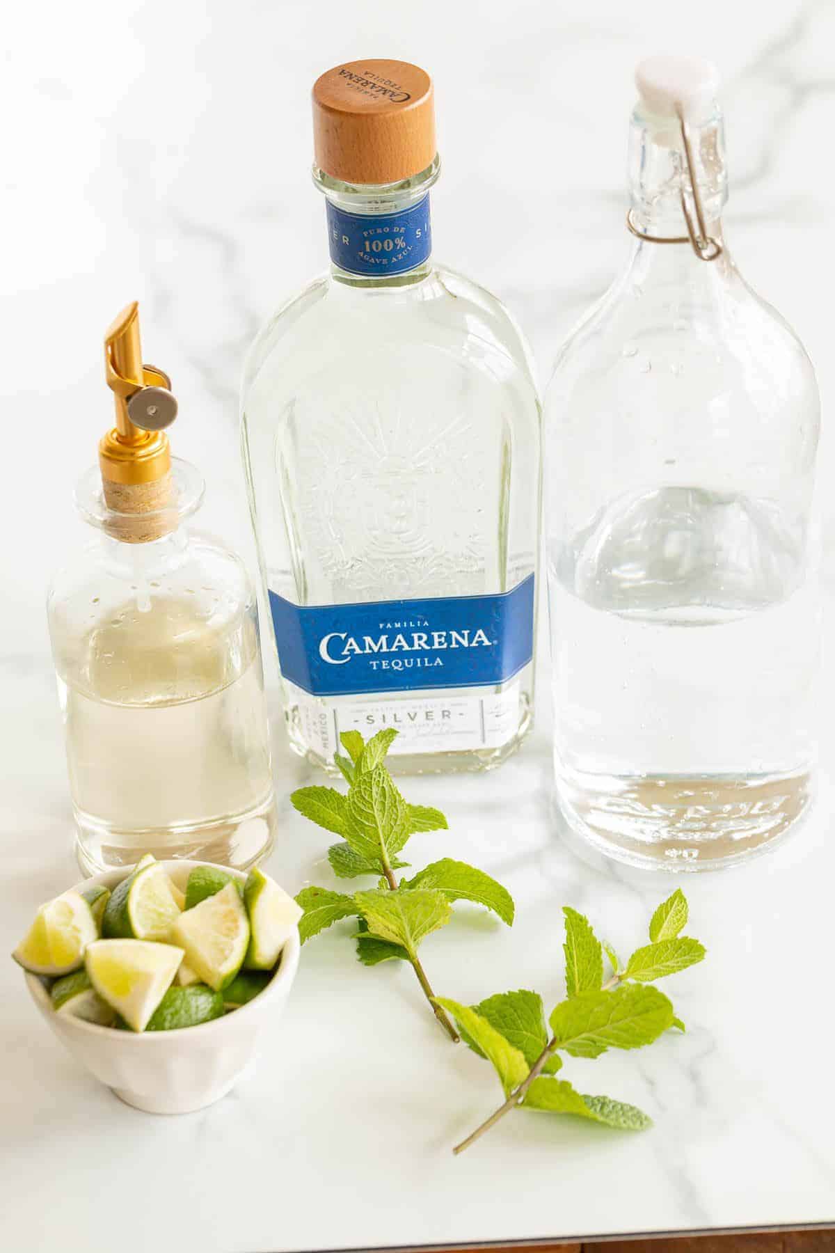 A display of clear bottles and ingredients for a tequila mojito on a white surface.