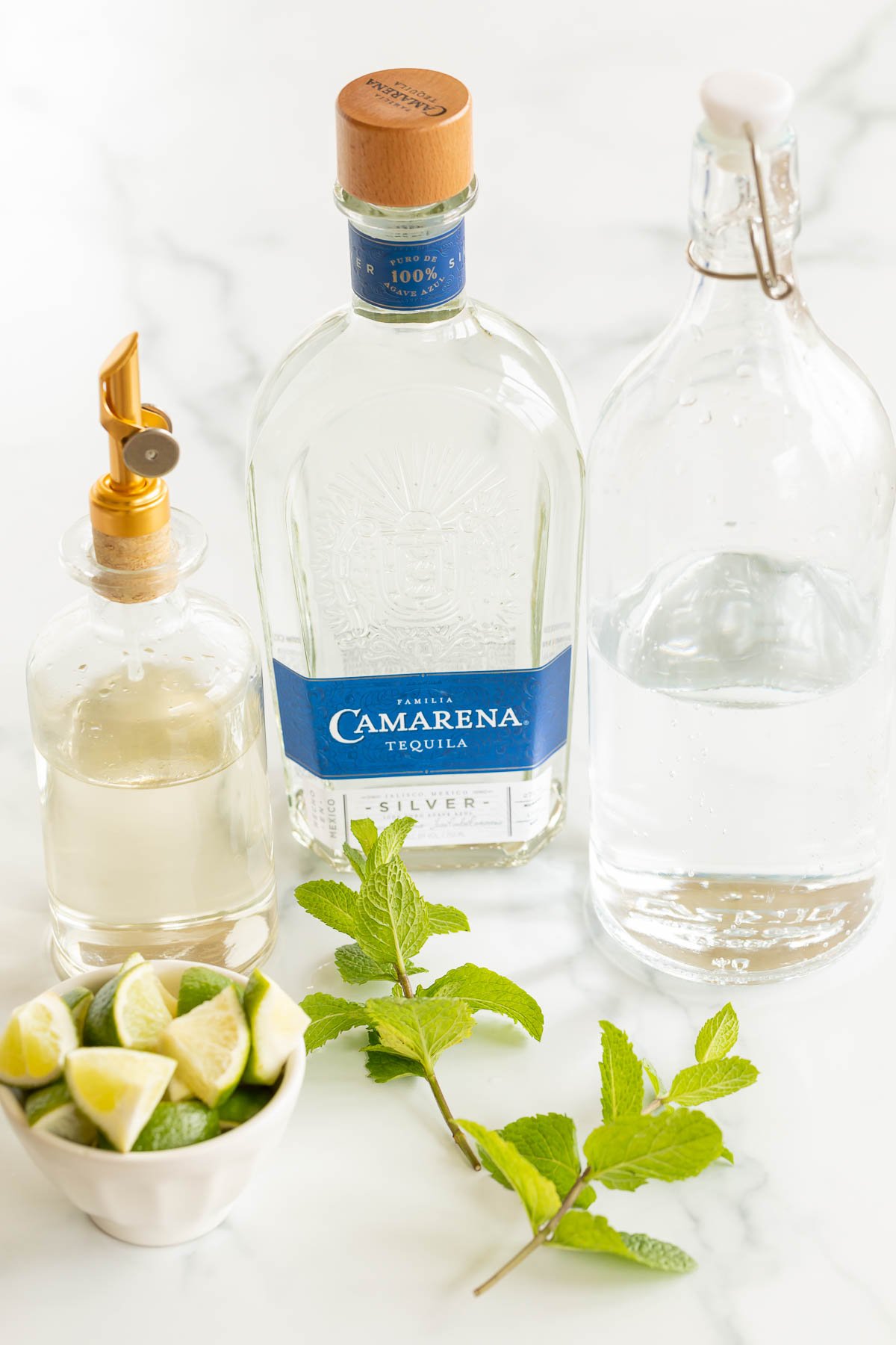 A bottle of Camarena silver tequila, a jug of clear liquid with a stopper, a dispenser with lime juice, and a bowl of lime wedges and mint leaves for a tequila mojito on a marble surface.