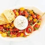 baked burrata bruschetta in a white oval bowl, gold spoon and crostini to the side.