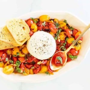 baked burrata bruschetta in a white oval bowl, golden spoon and crostini on the side.