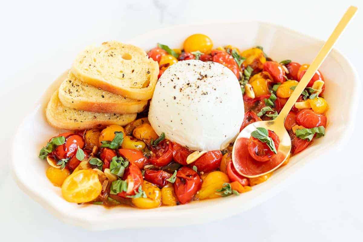 Burrata Bruschetta in a white serving bowl, crostini and a gold spoon to the side.