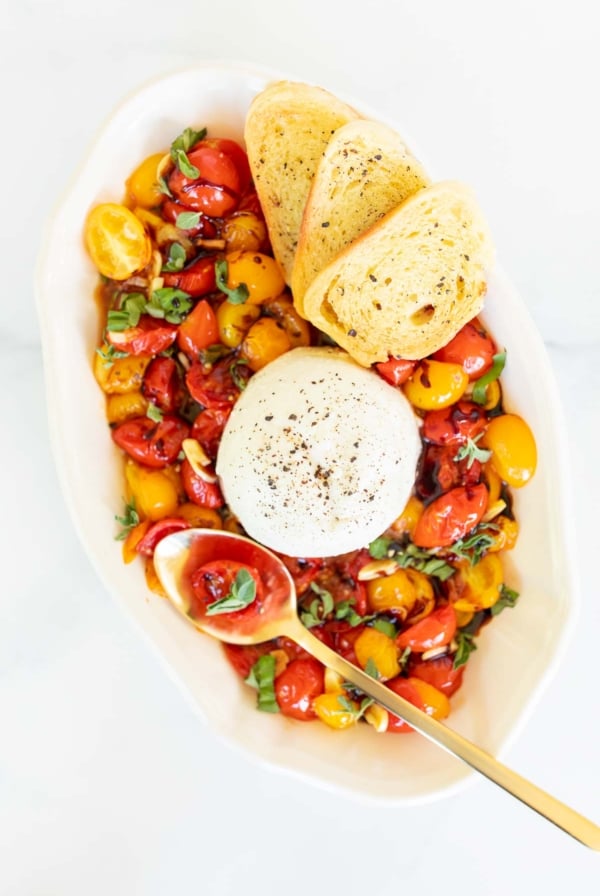 Burrata Bruschetta in a white serving bowl, crostini and a gold spoon to the side.