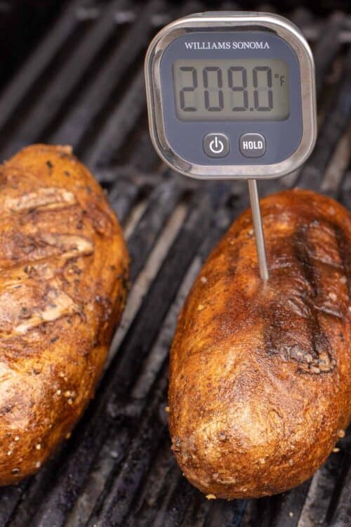 Grilled baked potatoes on a grill grate, one has a digital thermometer inside.