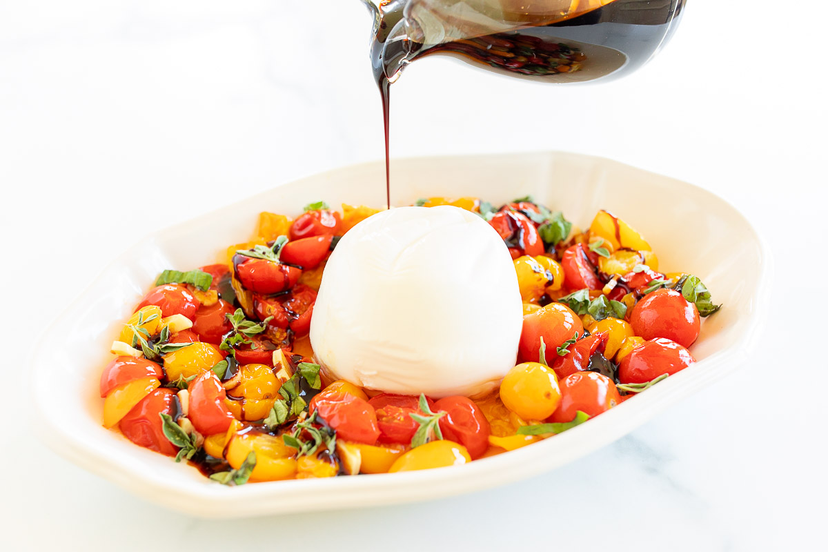 A small glass pitcher of balsamic glaze pouring over a dish of cherry tomatoes and mozzarella.
