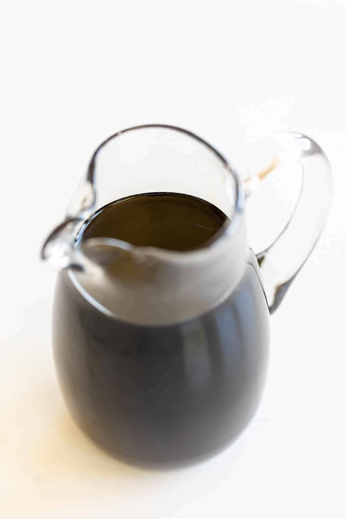 A clear pitcher full of balsamic glaze, on a white surface.