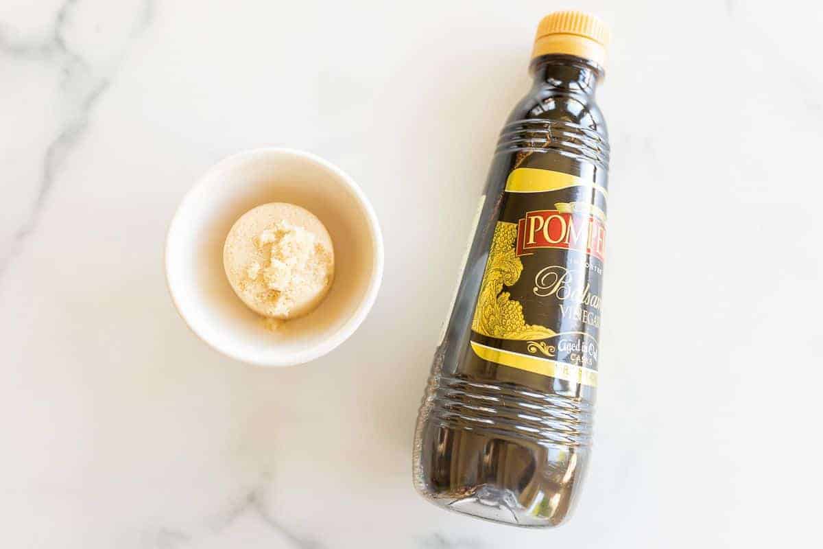 A bottle of balsamic vinegar and a small bowl of brown sugar on a marble surface.
