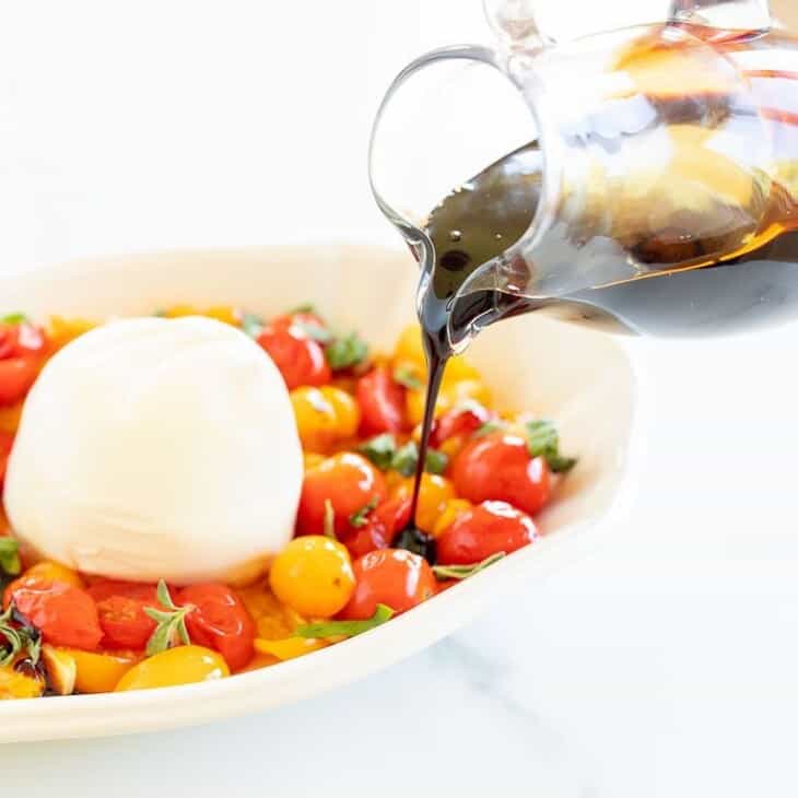 Baked burrata cheese surrounded by roasted cherry tomatoes, with a clear pitcher of balsamic glaze being drizzled over the top.