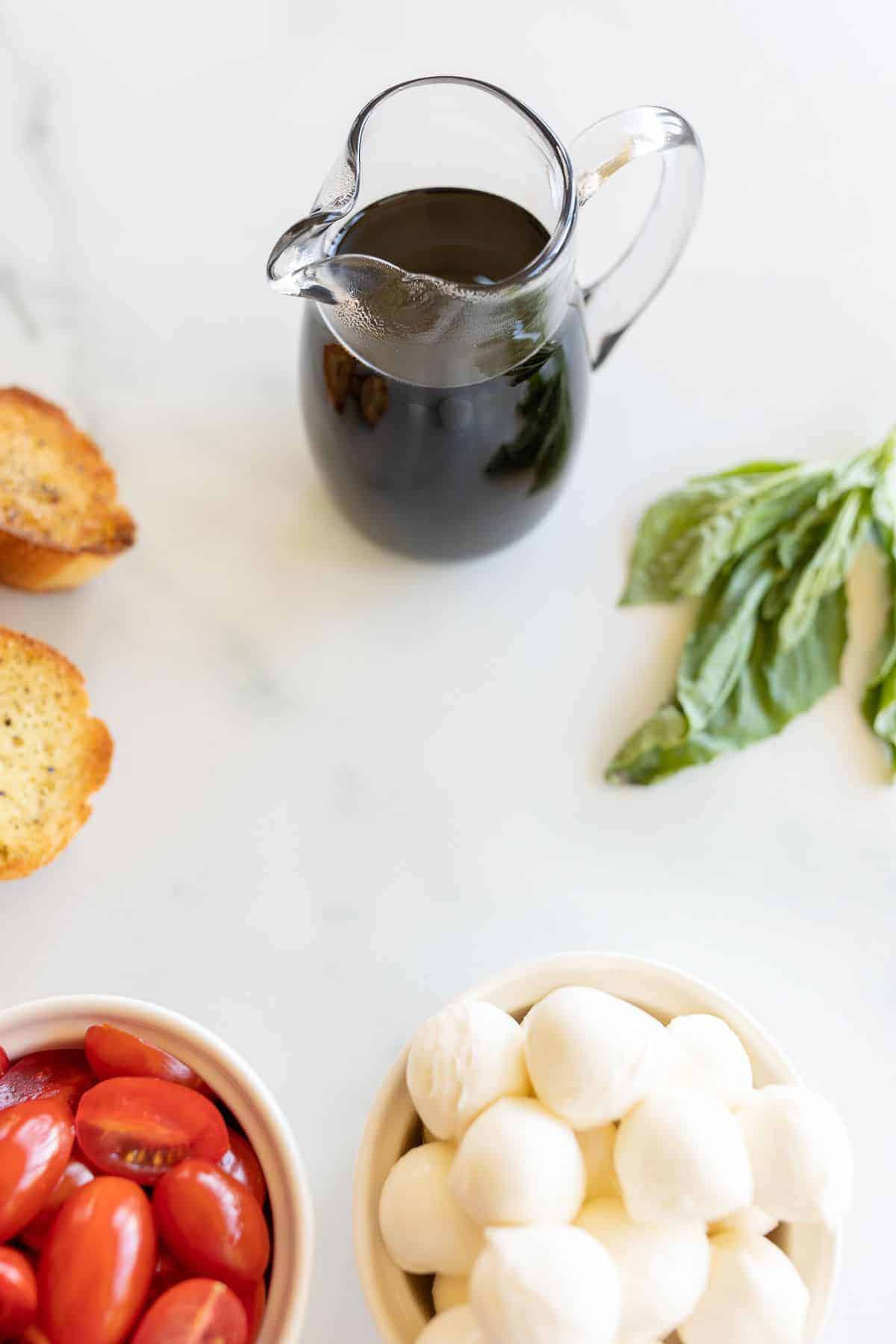 A small glass pitcher full of balsamic reduction, and bruschetta ingredients laid out on a marble surface.