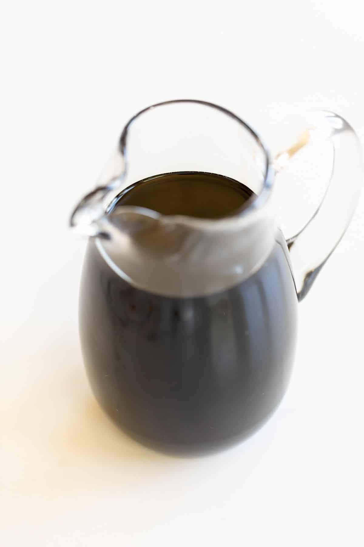 A clear pitcher full of balsamic glaze, on a white surface.