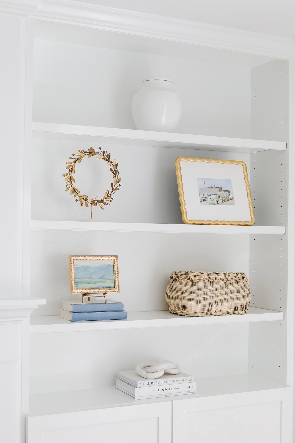 White built-in shelves showcase tasteful bookshelf decor, including a white vase, a gold wreath, a framed photo, a landscape painting, stacked books, a woven basket, and decorative beads.