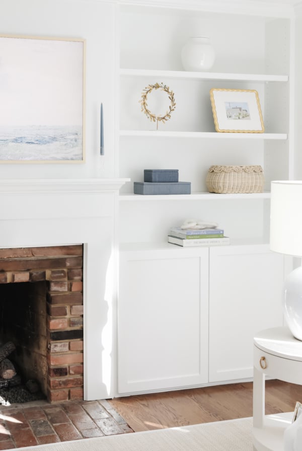 White built-in shelves showcase stylish bookshelf decor, including books, a basket, a framed picture, and a vase beside a brick fireplace in this bright, minimal living room.