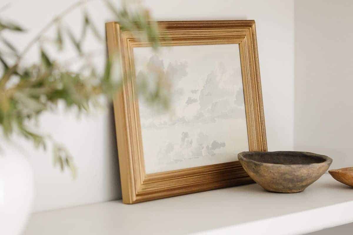 Bookshelf decor ideas of a gold framed piece of art and a small wood bowl in front, on white painted built ins.