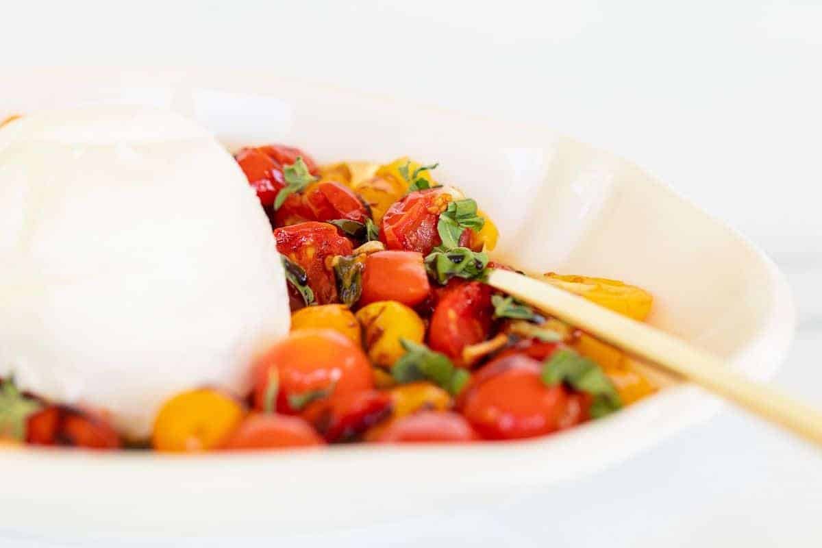A chunk of baked burrata cheese in a white bowl with roasted cherry tomatoes, gold spoon to the side.