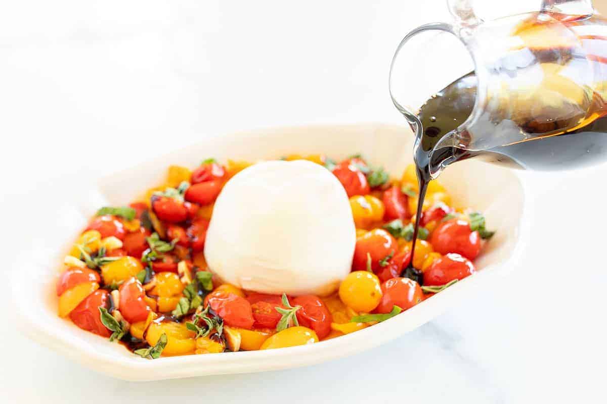 Roasted cherry tomatoes and basil topped with burrata cheese in a white serving dish, a hand drizzling balsamic over the top.
