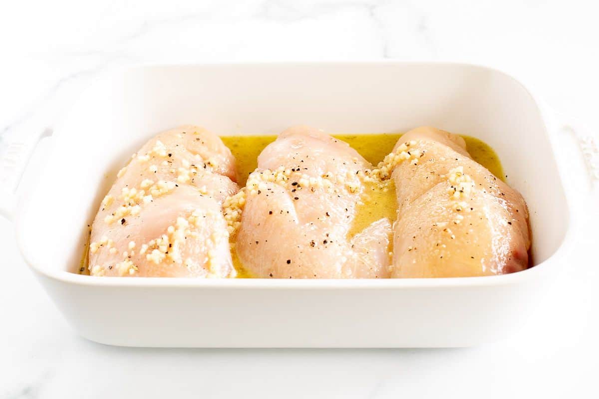 Raw chicken breasts topped with olive oil and seasonings in a white pan.