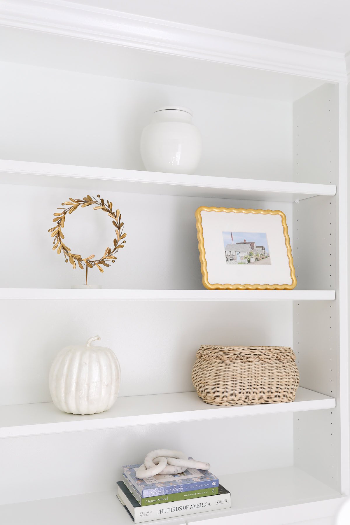 A white built-in shelf features stylish bookshelf decor including a white vase, gold wreath, framed picture, white pumpkin, wicker basket, coral accent piece, and stacked books.