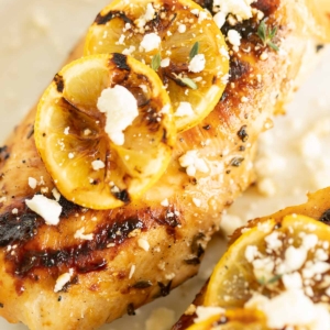 Greek chicken on a white surface, grilled lemon to the side.