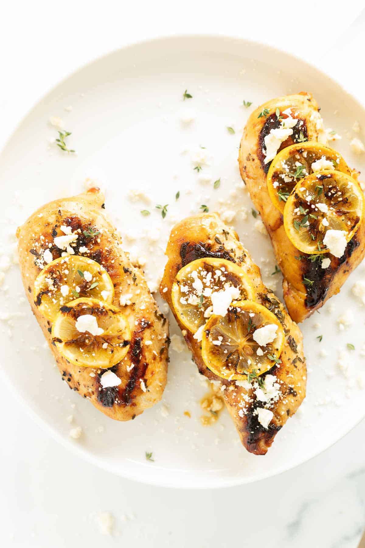 Greek chicken breast on a white plate, topped with grilled lemons and feta cheese and herbs.