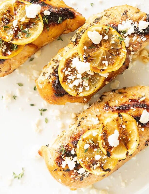 Greek chicken breast on a white plate, topped with grilled lemons and feta cheese and herbs.