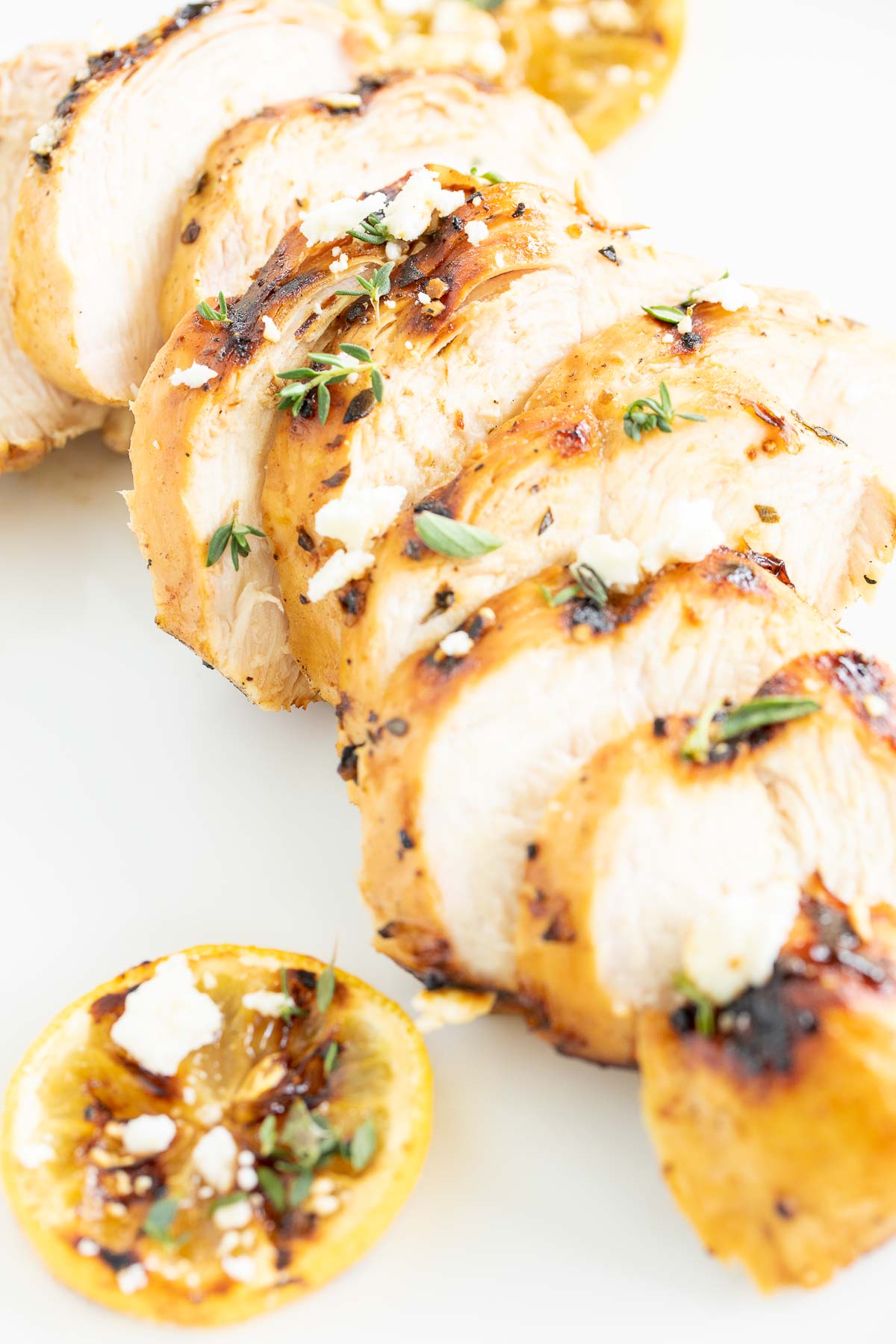 Slices of Greek marinated chicken on a white surface, grilled lemon to the side.