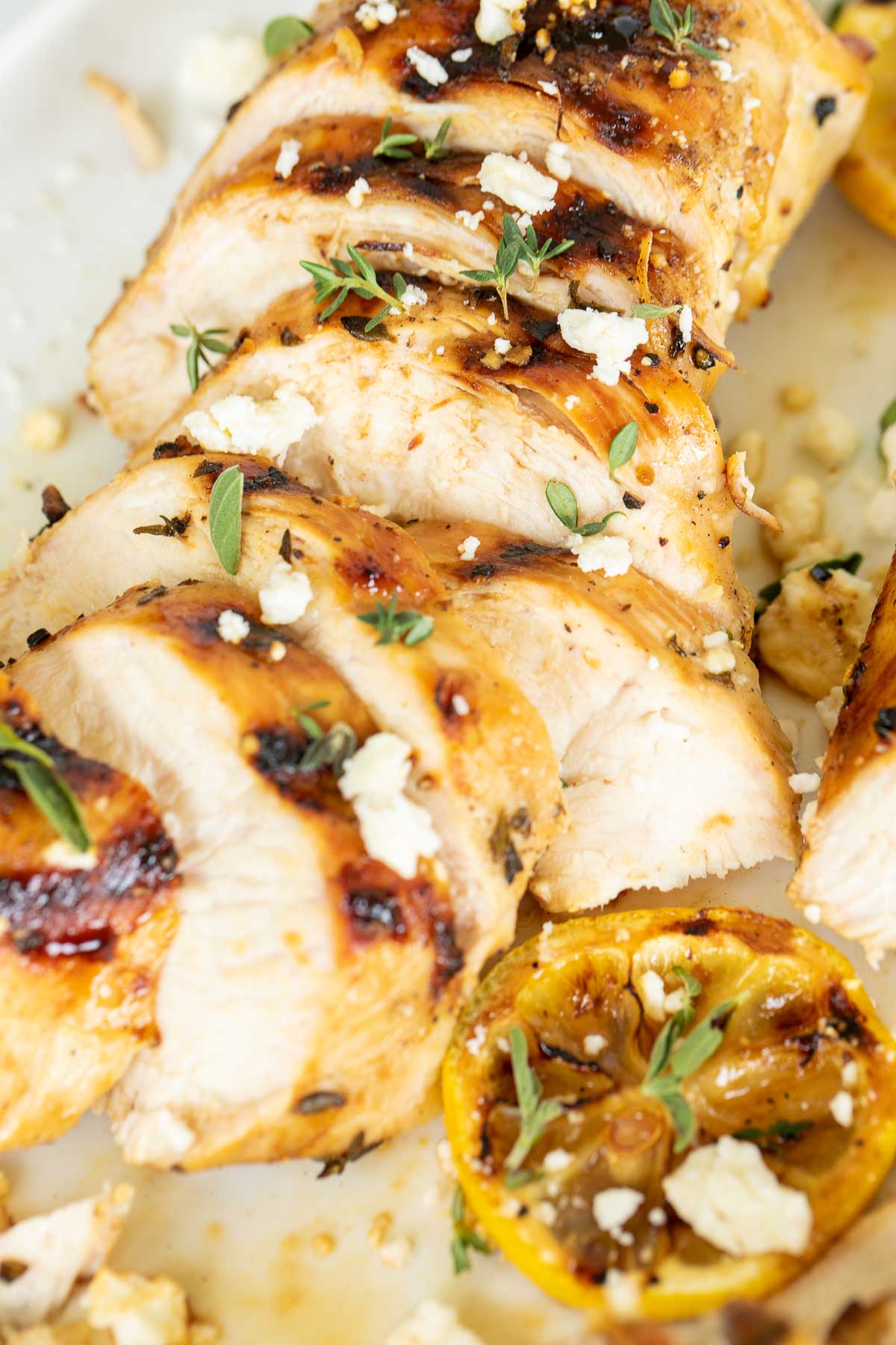 Slices of Greek marinated chicken on a white surface, grilled lemon to the side.