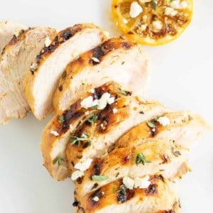 Slices of Greek marinated chicken on a white surface, grilled lemon on the side.