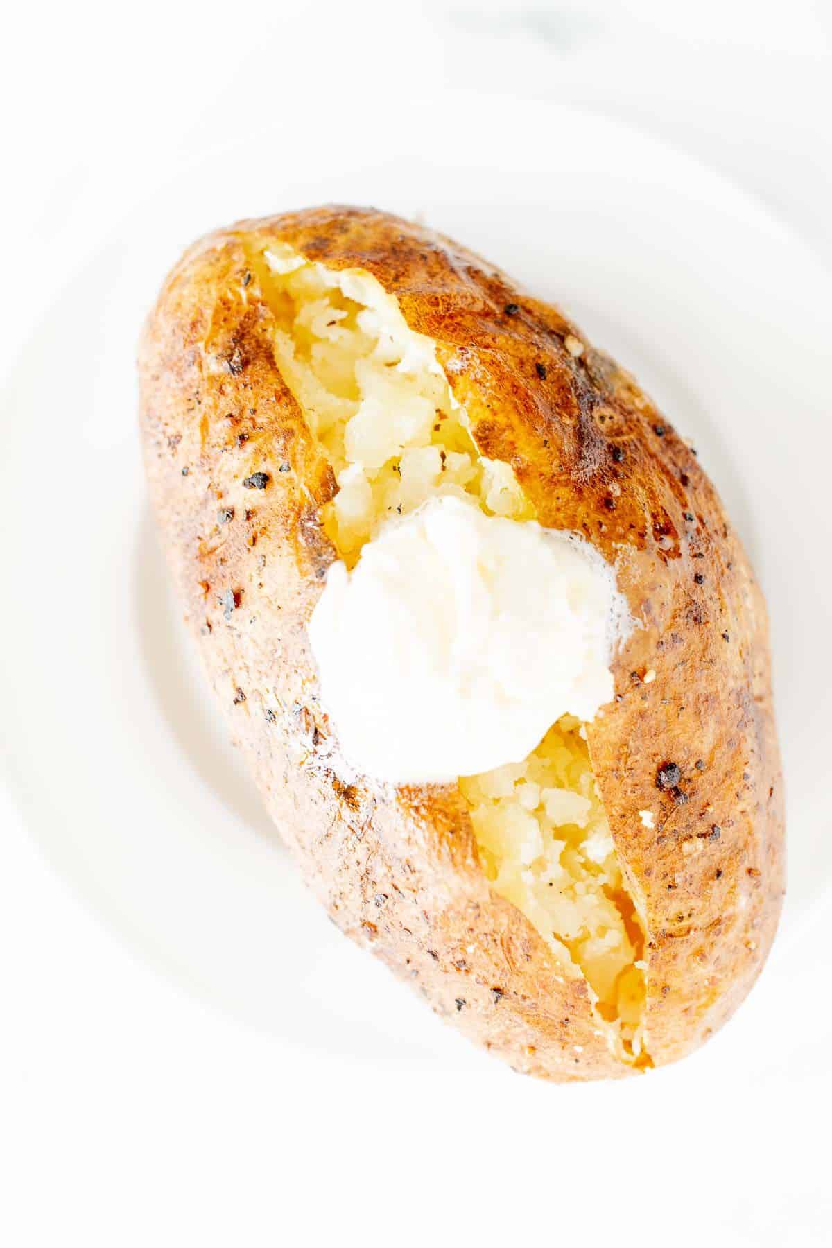 A single BBQ baked potato on a white plate, topped with butter and sour cream.