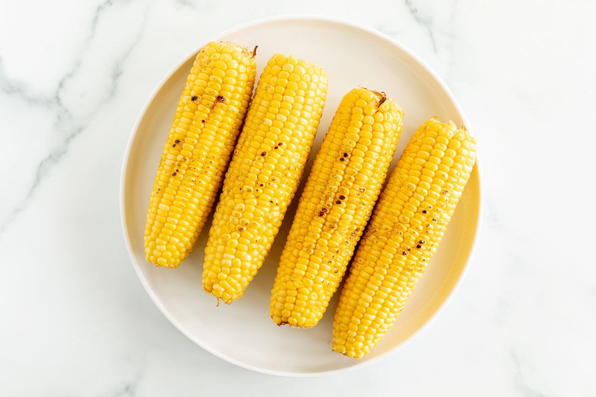 Four grilled corn on the cob on a white plate with a marble background.