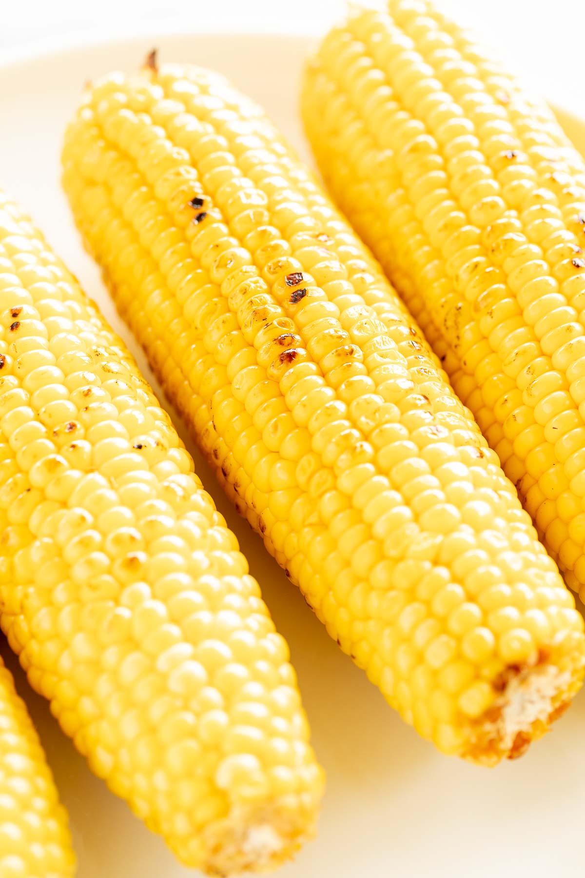 Three grilled corn on the cob on a white plate with char marks visible on the kernels.