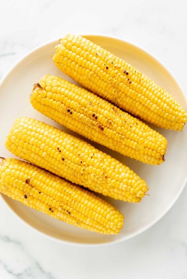 Four grilled corn on the cob with black pepper on a white oval plate against a marble background.