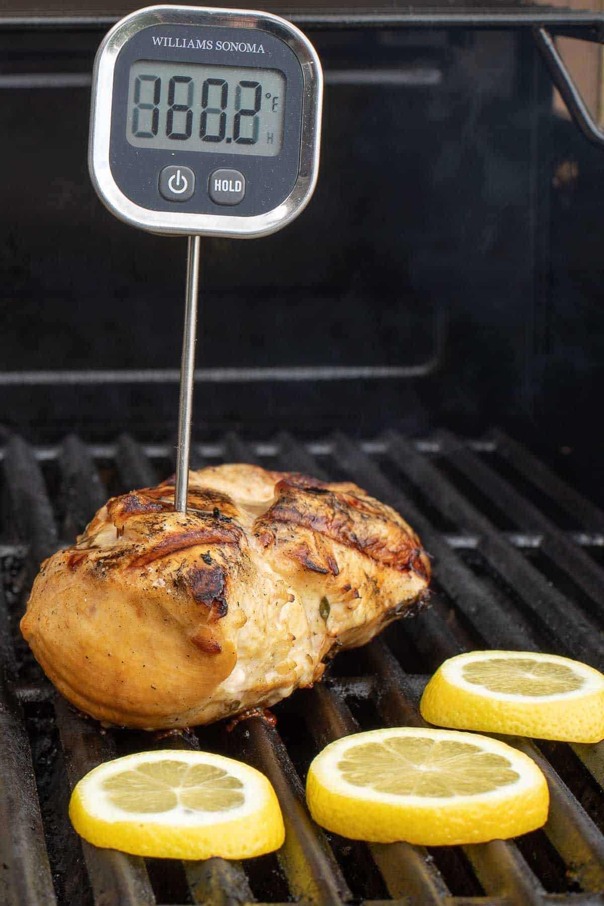 Greek chicken breast on a grill with a meat thermometer sticking out, slices of lemon being grilled too.