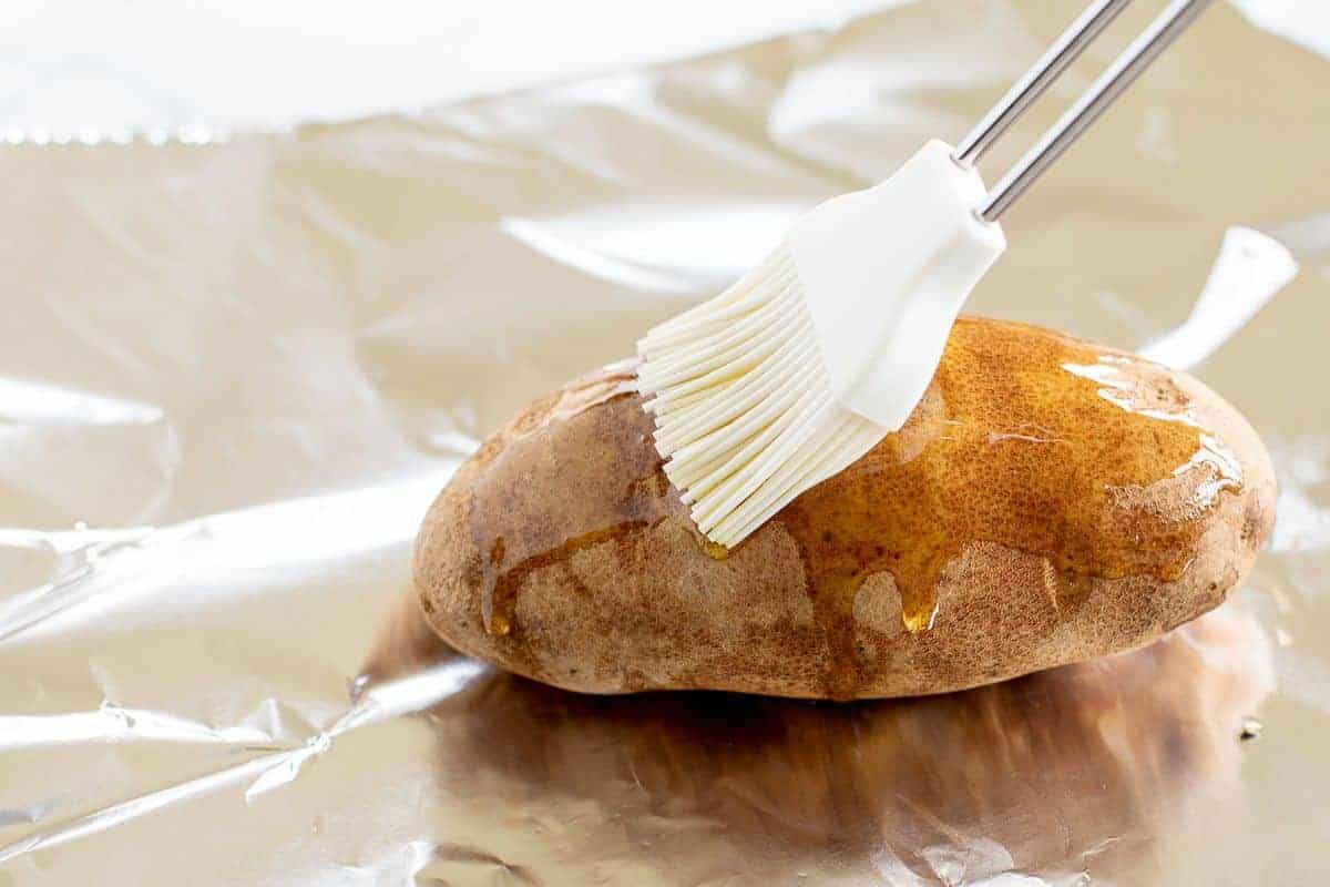 A russet potato on a sheet of foil, being brushed with olive oil.