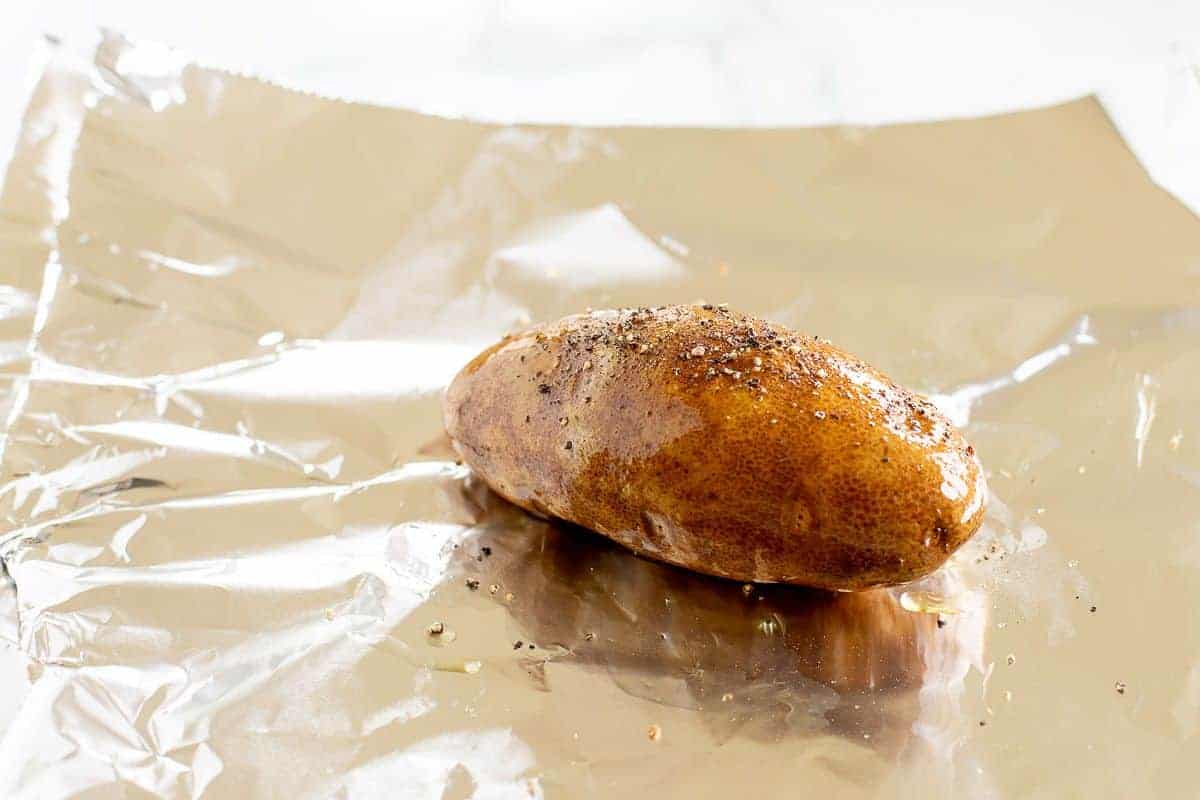 A single russet potato on a sheet of foil, topped with salt and pepper.