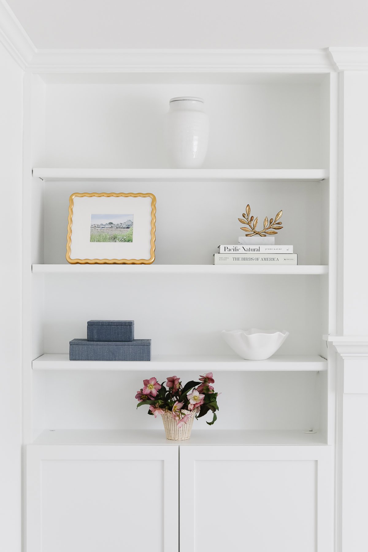 A white built-in shelf showcases stylish bookshelf decor, including a white vase, framed picture, books, a gold decorative object, stacked blue boxes, a white bowl, and a potted plant with pink flowers.