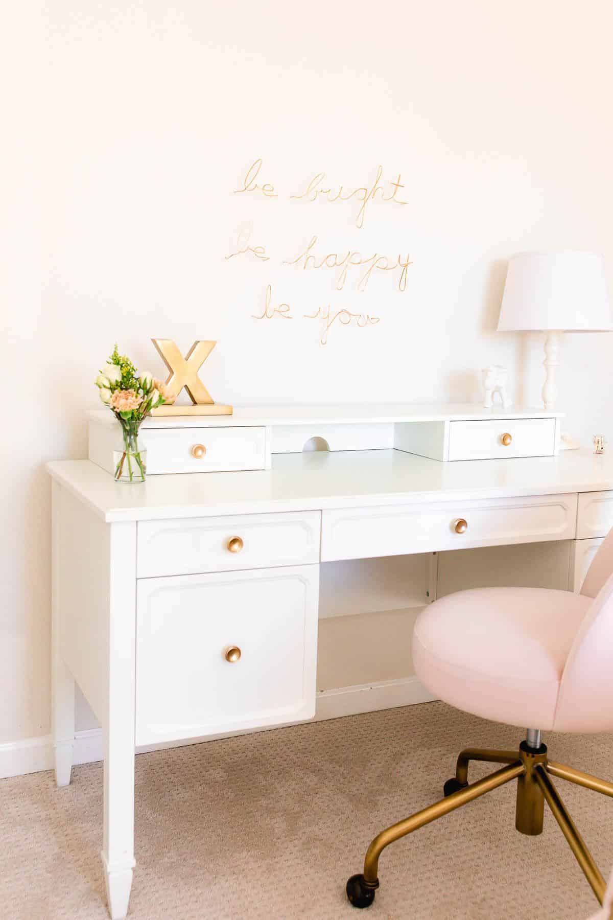 A white desk in a girl's bedroom with white walls and a pink chair.