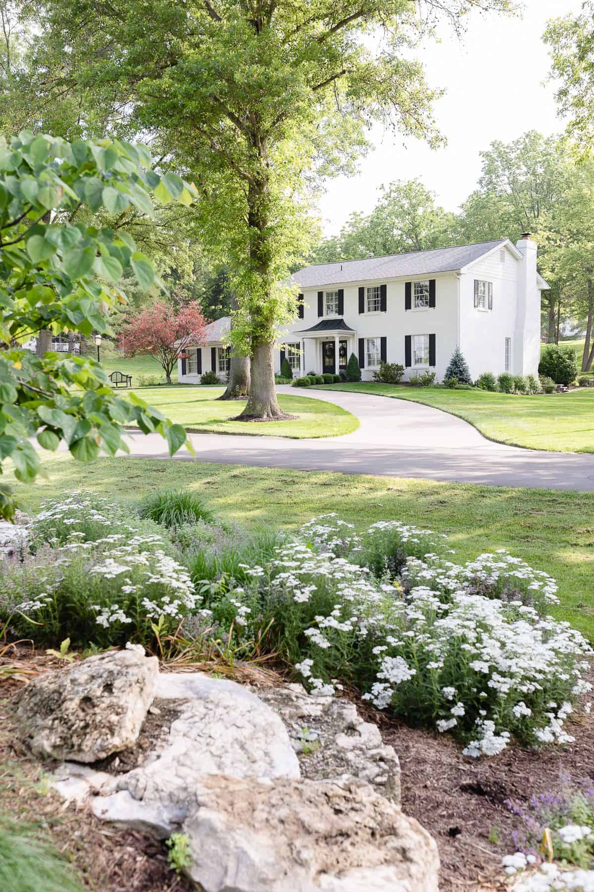 A white brick house sitting amongst green trees and flowers.