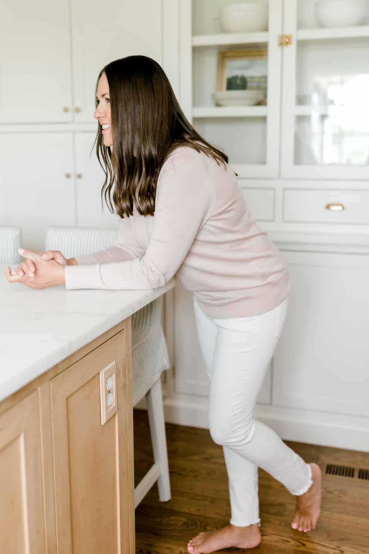 A brown haired woman leaning across a kitchen island in a white kitchen.